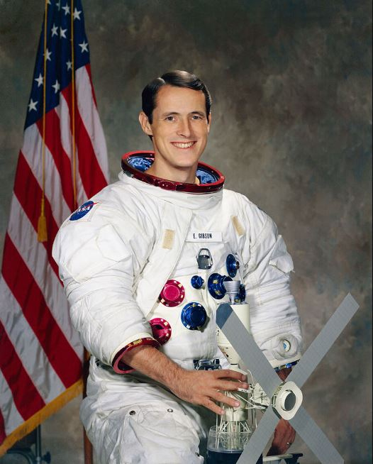 Photo of Edward Gibson holding a model in his white spacesuit.